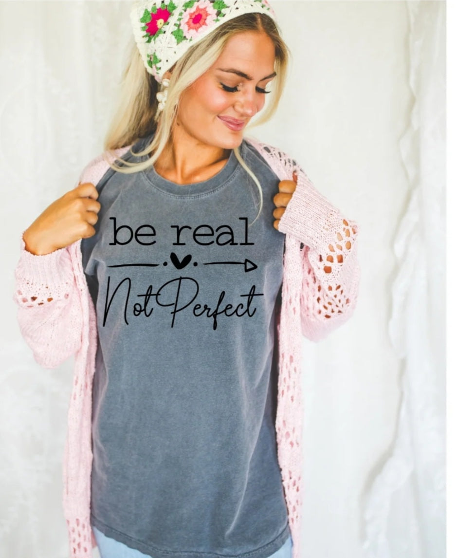 Woman wearing a gray t-shirt with 'be real not perfect' text, holding a pink lace garment.