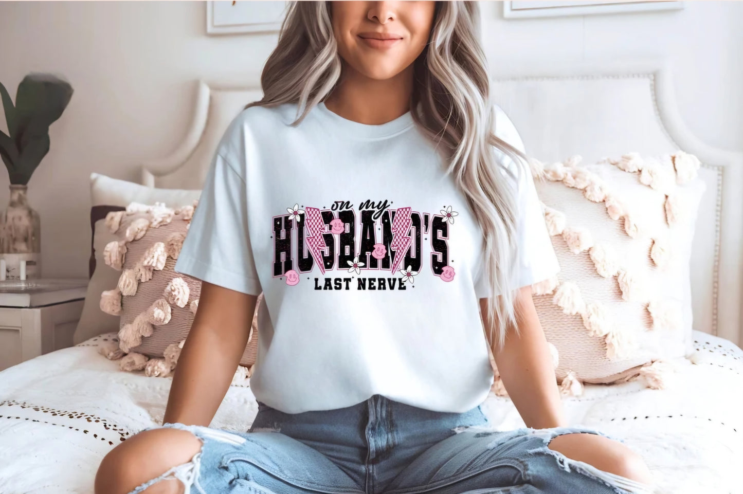 A woman sitting on a bed wearing a white t-shirt with the text 'On my husband's last nerve' printed on it.