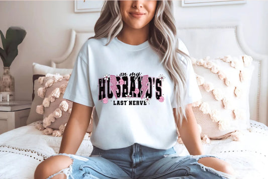 A woman sitting on a bed wearing a white t-shirt with the text 'On my husband's last nerve' printed on it.